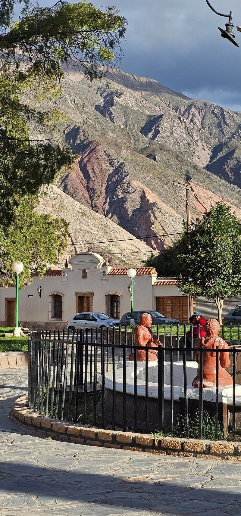 Fuente de la plaza con esculturas de terracota detrás de una reja, frente a una casa blanca y los cerros de colores.