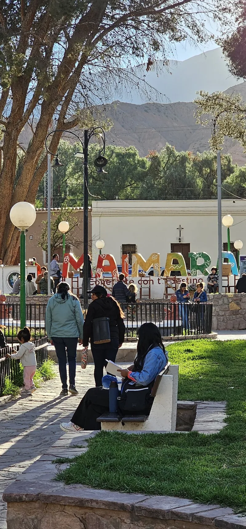 Vecinos caminando y descansando en la plaza, con el cartel de colores de Maimará y la cordillera al fondo.