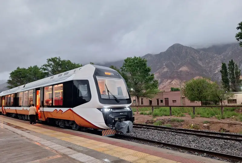 Tren moderno blanco y naranja con destino Volcán, detenido en el andén, con cerros nublados detrás.