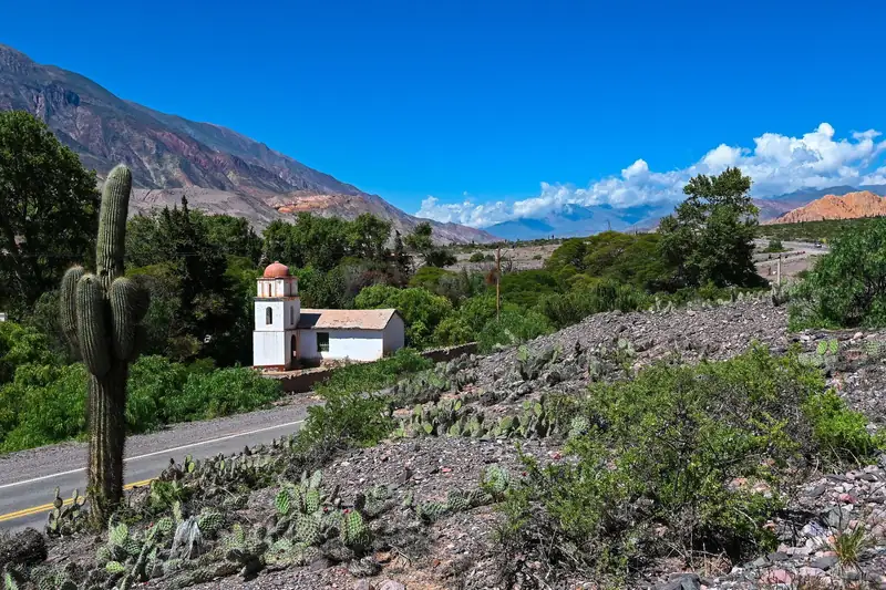 Capilla blanca con cúpula rojiza entre la vegetación, junto al camino, con un cardón y los cerros de colores.