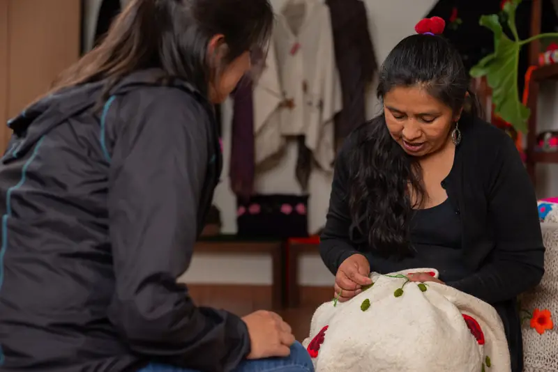 Una artesana borda con hilo verde y rojo un textil de lana blanco mientras otra persona la observa, con ponchos y cojines bordados detrás